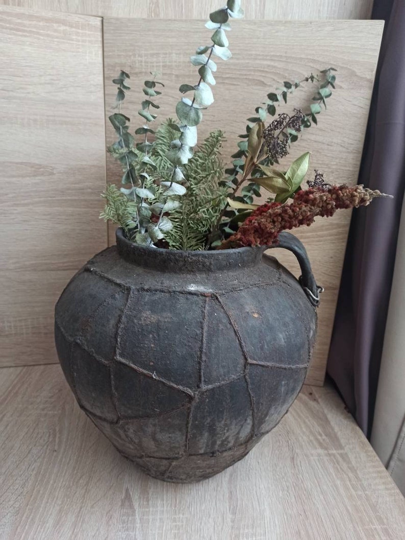 K&ouml;nnte beinhalten: Eine dunkelgraue Vase im antiken Stil mit einem einzigartigen geometrischen Muster, gef&uuml;llt mit getrocknetem Gr&uuml;n und Blumen. Die Vase hat einen Henkel und steht auf einer Holzoberfl&auml;che, was eine rustikale und nat&uuml;rliche &Auml;sthetik erzeugt.