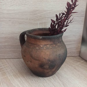 May include: A rustic brown clay pot with a handle, featuring a dark brown glaze and a rough texture. A sprig of dried burgundy-colored foliage is placed inside the pot.