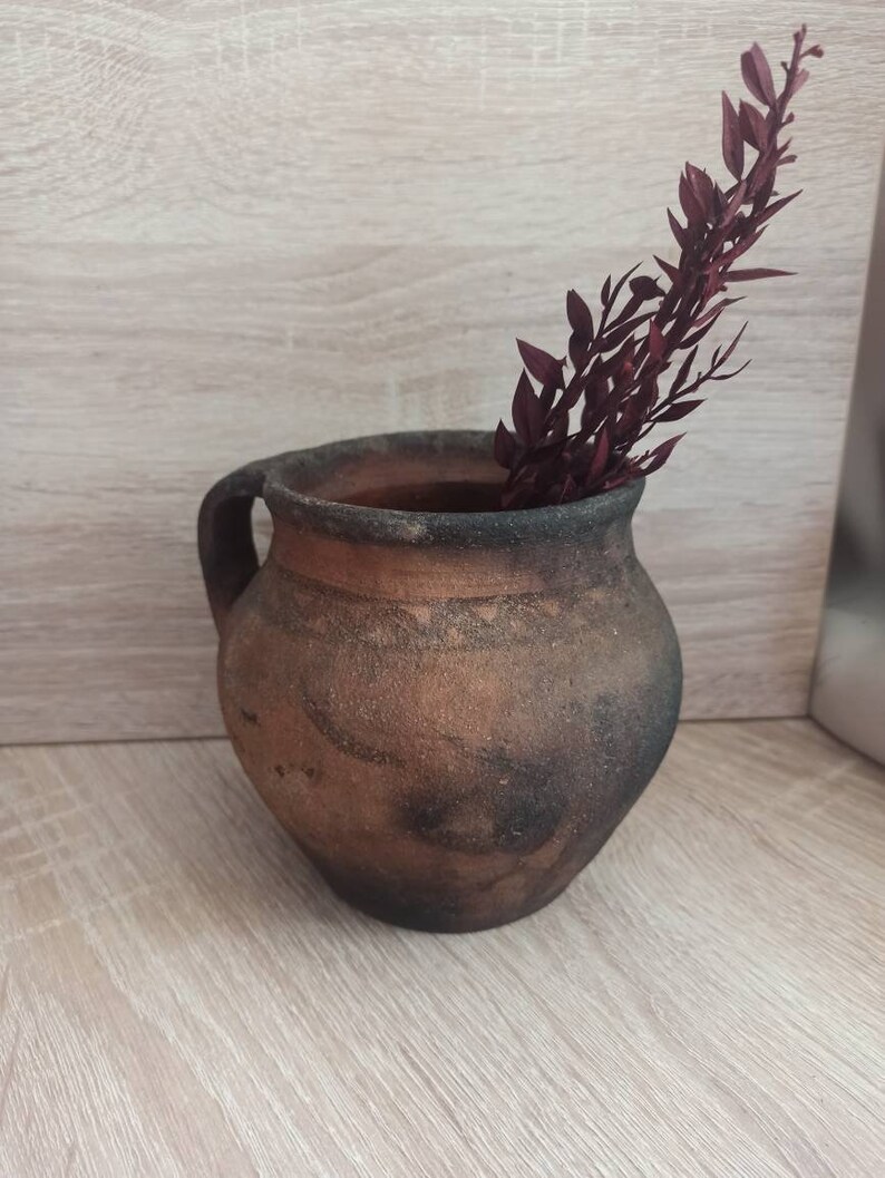 May include: A rustic brown clay pot with a handle, featuring a dark brown glaze and a rough texture. A sprig of dried burgundy-colored foliage is placed inside the pot.