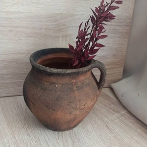 May include: A rustic brown clay pot with a handle, filled with a sprig of dried burgundy red foliage.
