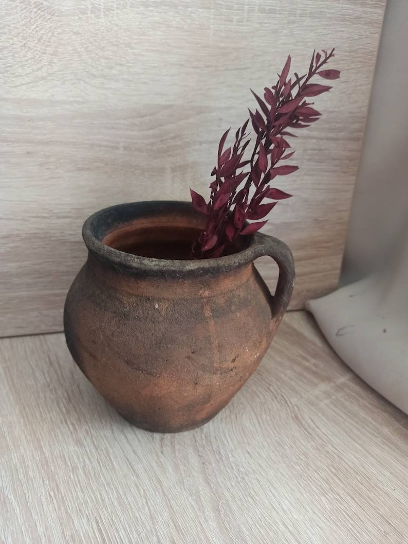 May include: A rustic brown clay pot with a handle, filled with a sprig of dried burgundy red foliage.
