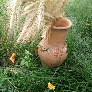 May include: A brown clay jug with green and orange floral designs sits in a field of green grass with dried wheat stalks inside.