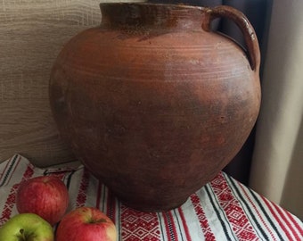 May include: A large, brown, earthenware pot with a handle sits on a white and red patterned tablecloth with three red apples and one green apple.