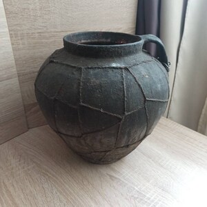 K&ouml;nnte beinhalten: Eine dunkle, runde Keramikkrug im antiken Stil mit Henkel. Der Krug hat eine dunkelgraue Farbe und ein geometrisches Muster aus Linien auf seiner Oberfl&auml;che. Der Krug steht auf einer hellen Holzoberfl&auml;che.