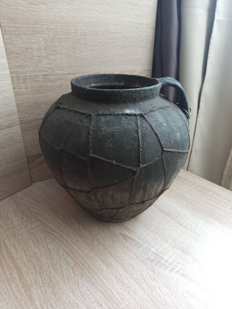 K&ouml;nnte beinhalten: Eine dunkle, runde Keramikkrug im antiken Stil mit Henkel. Der Krug hat eine dunkelgraue Farbe und ein geometrisches Muster aus Linien auf seiner Oberfl&auml;che. Der Krug steht auf einer hellen Holzoberfl&auml;che.