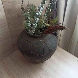 K&ouml;nnte beinhalten: Eine dunkelgraue, runde, strukturierte Vase mit geometrischem Muster, gef&uuml;llt mit getrocknetem Gr&uuml;n und Blumen. Das Arrangement umfasst Eukalyptus, Kiefer und andere getrocknete Elemente. Die Vase steht auf einer hellen Holzoberfl&auml;che.