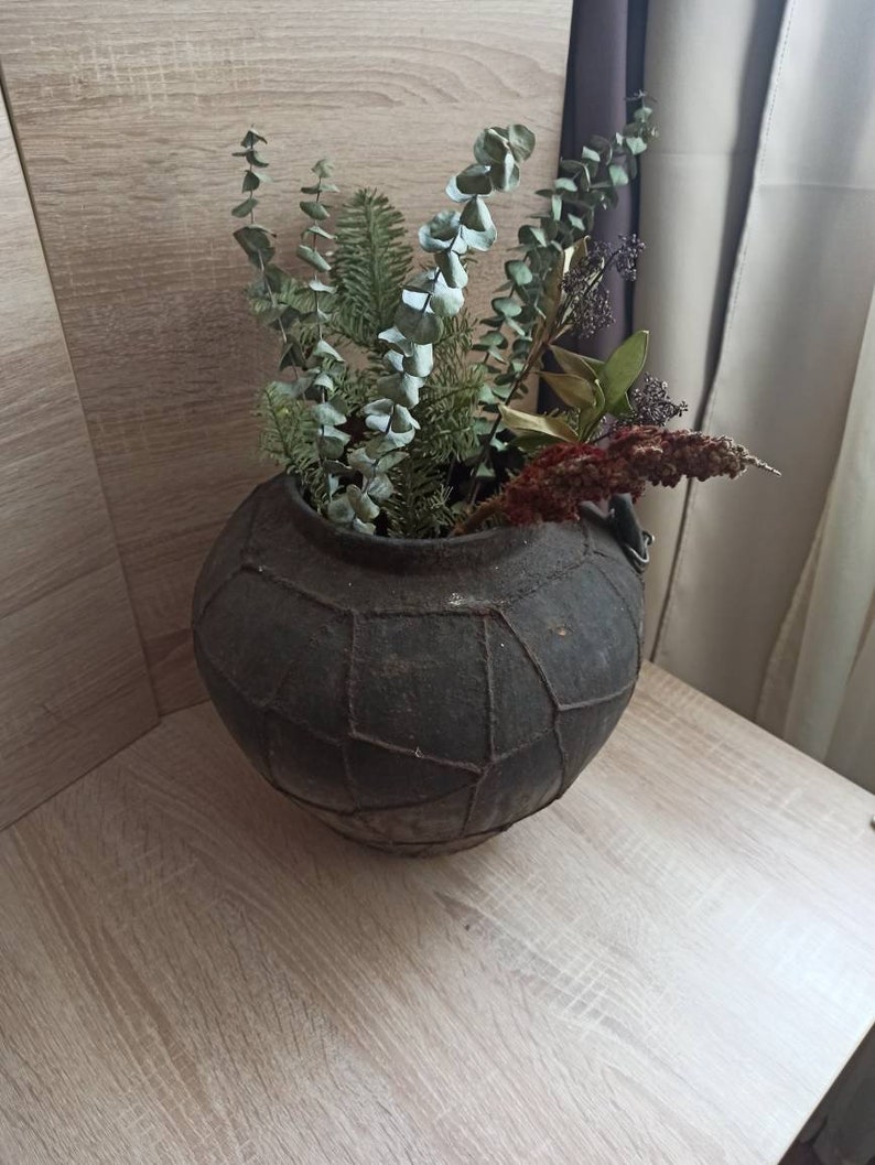 K&ouml;nnte beinhalten: Eine dunkelgraue, runde, strukturierte Vase mit geometrischem Muster, gef&uuml;llt mit getrocknetem Gr&uuml;n und Blumen. Das Arrangement umfasst Eukalyptus, Kiefer und andere getrocknete Elemente. Die Vase steht auf einer hellen Holzoberfl&auml;che.