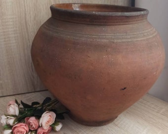 May include: A large, brown, earthenware pot with a rounded body and a wide opening. The pot has a rough, textured surface and appears to be handmade.