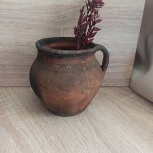 May include: A rustic brown clay pot with a dark brown rim and a handle on one side. A sprig of dried burgundy red flowers is inside the pot.