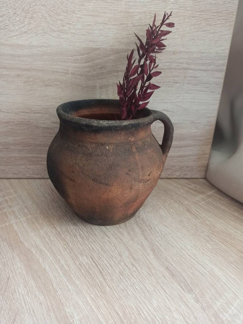 May include: A rustic brown clay pot with a dark brown rim and a handle on one side. A sprig of dried burgundy red flowers is inside the pot.