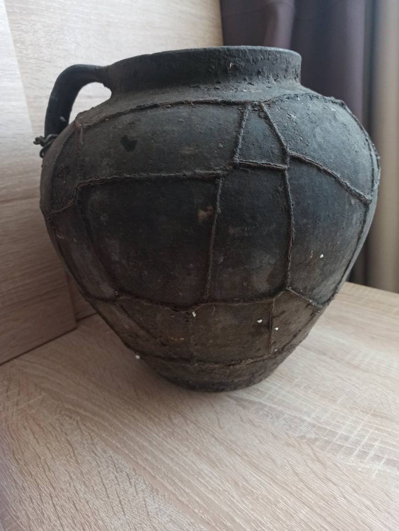 May include: A large, dark brown clay pot with a wide mouth and a handle. The pot is decorated with a pattern of woven strips.