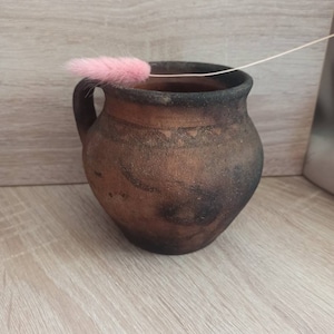 May include: A brown clay pot with a handle, sitting on a light wood surface. A pink feather is resting on the rim of the pot.