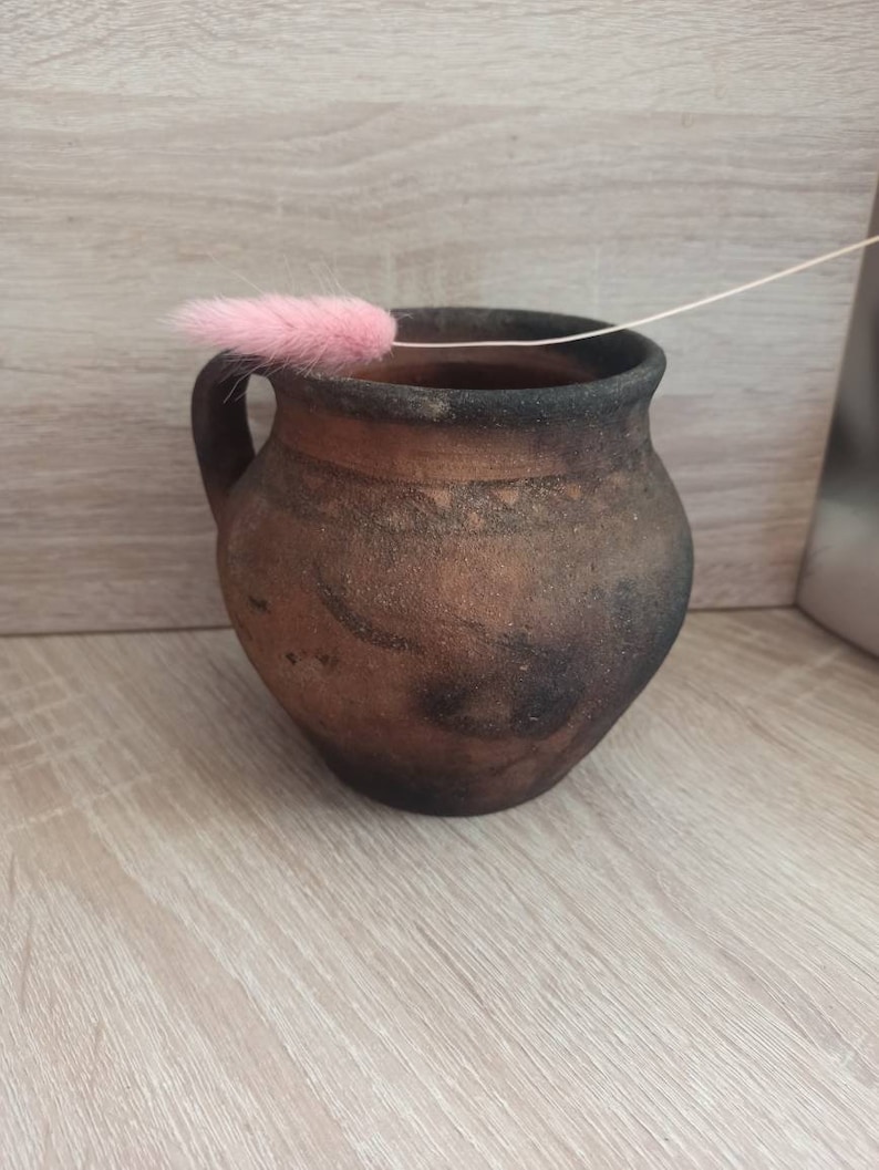 May include: A brown clay pot with a handle, sitting on a light wood surface. A pink feather is resting on the rim of the pot.