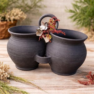Puede incluir: Dos jarrones de cerámica negra conectados, con asa y líneas grabadas decorativas. Los jarrones están adornados con flores secas y se asientan sobre una superficie de madera. Los jarrones tienen un aspecto rústico y artesanal.