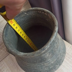 Pode incluir: Um pote de cer&acirc;mica cinza envelhecido com uma al&ccedil;a. A abertura interna mede aproximadamente 18 cm de di&acirc;metro. O pote est&aacute; sobre uma superf&iacute;cie de madeira clara.