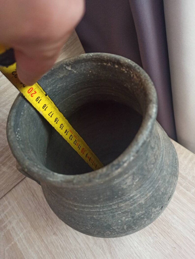 Pode incluir: Um pote de cer&acirc;mica cinza envelhecido com uma al&ccedil;a. A abertura interna mede aproximadamente 18 cm de di&acirc;metro. O pote est&aacute; sobre uma superf&iacute;cie de madeira clara.