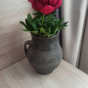 Pode incluir: Um vaso de cer&acirc;mica escuro e r&uacute;stico cont&eacute;m uma vibrante flor de pe&ocirc;nia vermelha com folhas verdes. O vaso tem uma al&ccedil;a e fica em uma superf&iacute;cie de madeira clara. A flor est&aacute; em plena flora&ccedil;&atilde;o.