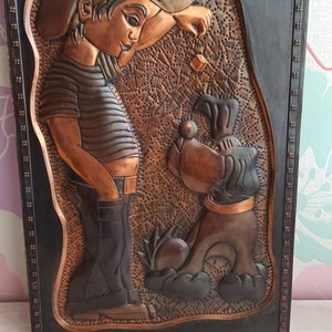May include: A copper wall art piece depicting a boy in a cap and a dog. The boy is holding a small object in his hand and the dog is looking up at him. The artwork is framed in a black frame with a decorative border.