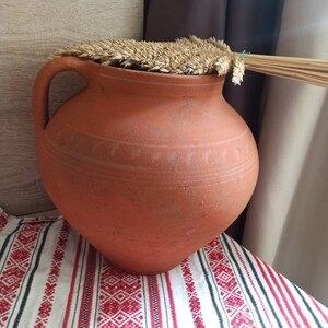 May include: A large, terracotta-colored clay pot with a wide mouth and a handle. The pot is covered with a layer of dried wheat stalks.