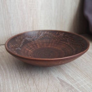 May include: A brown ceramic bowl with a decorative design of leaves around the rim. The bowl is slightly curved and has a smooth, glossy finish.