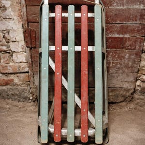 Può includere: Una slitta in legno vintage con telaio in metallo. La slitta presenta doghe verticali rosse e verde acqua alternate. Una corda è attaccata in alto. La slitta è probabilmente utilizzata per lo svago invernale.