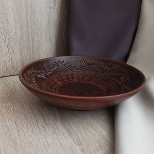 May include: A brown ceramic bowl with a decorative floral design around the rim. The bowl is sitting on a light brown wooden surface.