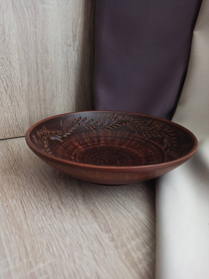 May include: A brown ceramic bowl with a decorative floral design around the rim. The bowl is sitting on a light brown wooden surface.