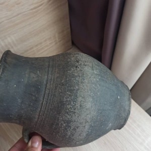 Pode incluir: Um jarro de cer&acirc;mica cinza escuro, de estilo antigo, com uma al&ccedil;a. O jarro tem um corpo arredondado e um pesco&ccedil;o estreito. A superf&iacute;cie tem uma apar&ecirc;ncia texturizada e envelhecida, sugerindo que &eacute; uma pe&ccedil;a antiga. O jarro &eacute; segurado por uma m&atilde;o.