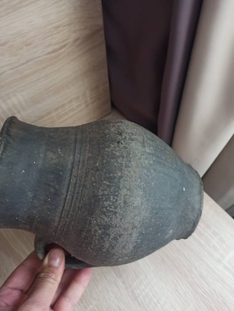 Pode incluir: Um jarro de cer&acirc;mica cinza escuro, de estilo antigo, com uma al&ccedil;a. O jarro tem um corpo arredondado e um pesco&ccedil;o estreito. A superf&iacute;cie tem uma apar&ecirc;ncia texturizada e envelhecida, sugerindo que &eacute; uma pe&ccedil;a antiga. O jarro &eacute; segurado por uma m&atilde;o.