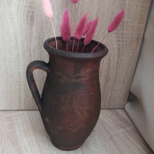 May include: A brown ceramic pitcher with a handle, filled with pink and light pink dried flowers.