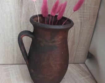 Pode incluir: Um jarro de cerâmica marrom com alça, cheio de flores secas rosa e rosa claro.