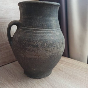Pode incluir: Jarro de cer&acirc;mica cinza escuro em estilo antigo com uma al&ccedil;a. O jarro tem uma base larga, um corpo arredondado e uma borda alargada. A superf&iacute;cie tem uma apar&ecirc;ncia texturizada e envelhecida, sugerindo que &eacute; vintage ou feito &agrave; m&atilde;o.