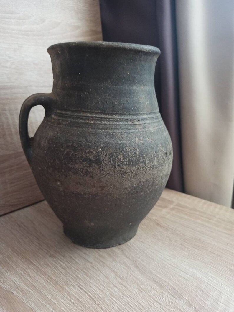 Pode incluir: Jarro de cer&acirc;mica cinza escuro em estilo antigo com uma al&ccedil;a. O jarro tem uma base larga, um corpo arredondado e uma borda alargada. A superf&iacute;cie tem uma apar&ecirc;ncia texturizada e envelhecida, sugerindo que &eacute; vintage ou feito &agrave; m&atilde;o.