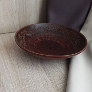 May include: A brown ceramic bowl with a decorative floral design. The bowl is empty and sitting on a light wood surface.