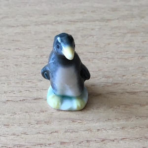 May include: A small ceramic figurine of a penguin with a dark grey back, a white chest, and a pale yellow beak. The penguin is standing on a white base with subtle green shading, featuring a smooth, glossy finish and a rounded, upright posture.