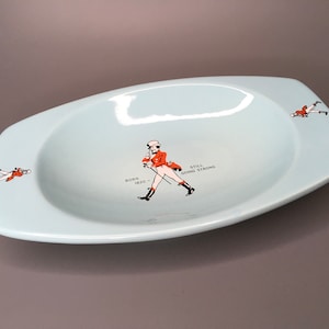 May include: A light blue ceramic ashtray featuring a red and black illustration of a man in a red coat and a top hat. The text "BORN 1820 - STILL GOING STRONG" is printed on the ashtray.