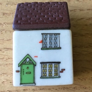 May include: A small rectangular eraser shaped like a house with a textured brown roof. The white body features a printed illustration of a green front door with a peaked roof, two multi-pane windows, and small orange brick-like accents.
