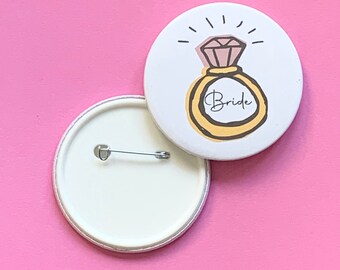 May include: White button with a gold ring and pink diamond design. The ring has the word "Bride" written on it. The back of the button is white with a safety pin.