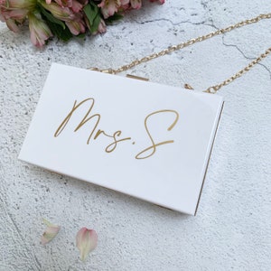 Puede incluir: Clutch blanca con una correa de cadena dorada. La cartera tiene el texto "Mrs. S" escrito en letras doradas.