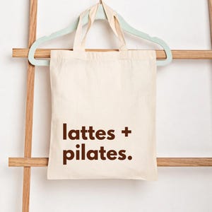 Puede incluir: Bolso tote de color crema con las palabras "lattes + pilates." impresas en marr&oacute;n. La bolsa est&aacute; colgada de una percha verde claro, que descansa sobre una escalera de madera.