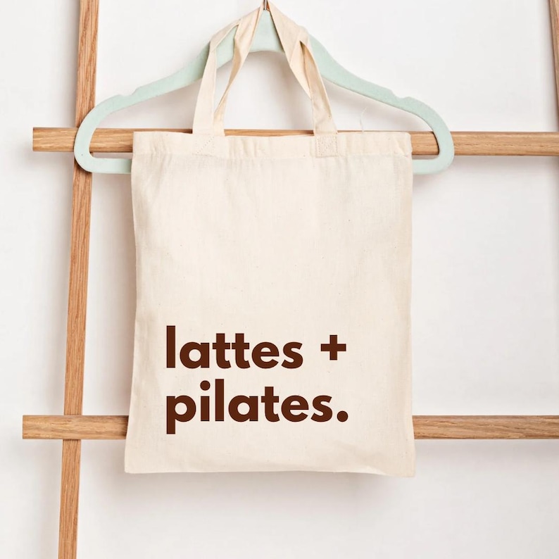 Puede incluir: Bolso tote de color crema con las palabras "lattes + pilates." impresas en marr&oacute;n. La bolsa est&aacute; colgada de una percha verde claro, que descansa sobre una escalera de madera.