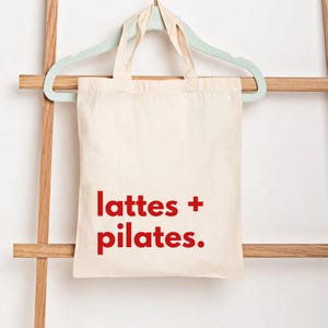 Puede incluir: Bolso tote de color crema con texto rojo "lattes + pilates." La bolsa est&aacute; colgada de una percha verde claro, que descansa sobre una estructura de madera en forma de escalera. La bolsa tiene dos asas.