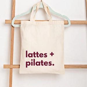 Puede incluir: Bolso tote de color crema con las palabras "lattes + pilates." impresas en un color burdeos intenso. La bolsa est&aacute; colgada de una percha verde claro, que descansa sobre una escalera de madera.