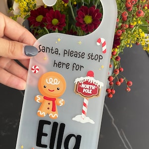 May include: A frosted plastic door hanger with a festive Christmas design. It features a gingerbread man, a North Pole sign, and the name "Ella". The text reads "Santa, Please stop here for". Candy and a Christmas tree are also included.