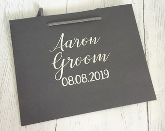 May include: A gray gift bag with a white rope handle. The bag is personalized with the text "Aaron Groom 08.08.2019" in white.