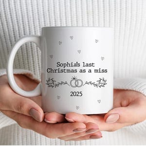Puede incluir: Taza de cerámica blanca con el texto "Sophia's last Christmas as a miss 2025" en negro. La taza presenta un diseño de corazón y anillos. La taza está sostenida por una persona que lleva un suéter blanco.