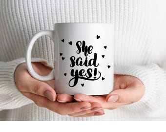Op de afbeelding: Witte keramische mok met de tekst "She said yes!" in zwart script, omringd door kleine zwarte hartvormen. De mok heeft een gebogen handvat en wordt vastgehouden door een persoon die een witte trui draagt.