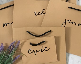 May include: Four brown paper gift bags with black handles and names written on them. The names are "Rebekah", "Jennie", "Evie", and "M".