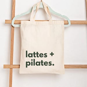 Puede incluir: Bolso tote de color crema con el texto "lattes + pilates." en verde oscuro. La bolsa est&aacute; colgada de una percha verde claro, que descansa sobre una estructura de madera en forma de escalera. La bolsa tiene dos asas.
