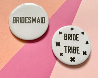 May include: Two white round buttons with black text. One button says "BRIDESMAID" and the other button says "BRIDE TRIBE" with black crosses around the text.